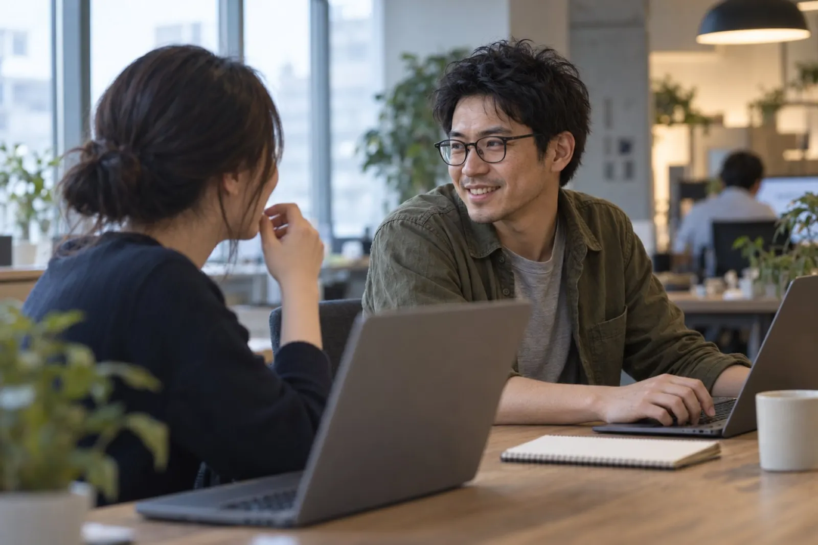 オフィスの長い机で、隣の席の同僚と話しながら笑う石田（イメージ・見本）